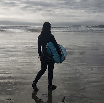 A picture of Emma Ritchie - Life and Wellness Coach walking away from the camera wearing a wetsuit and carrying a surfboard