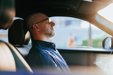 Bald man in glasses sits in a car, eyes closed, wearing a blue shirt, looking uncertain and stressed.