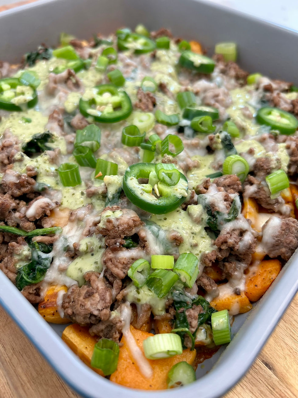 Eye-level view of a baked dish with ground beef, spinach, sweet potatoes, and melted cheese topped with creamy avocado drizzle