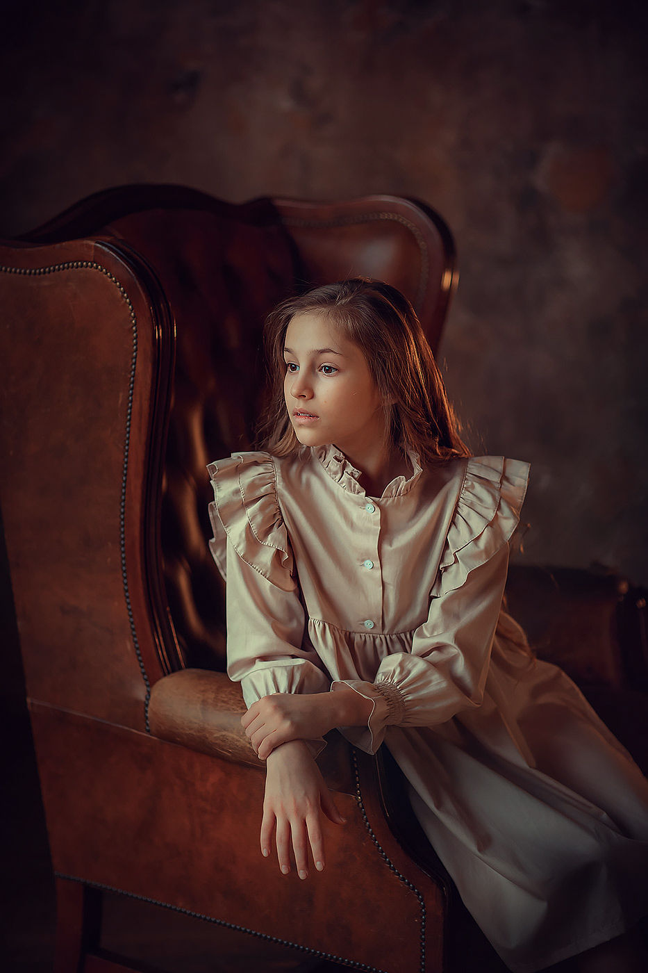 Girl in beige dress sitting on the brown chair in the dark studio