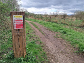 Wooden signpost in nature reserve warns of deep cold water and prohibits activities. Dirt path and cloudy sky in the background.
