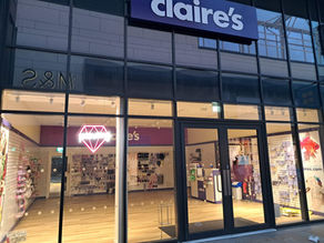 Storefront of Claire's with large glass windows displaying jewelry and accessories. A pink neon diamond light is on inside.