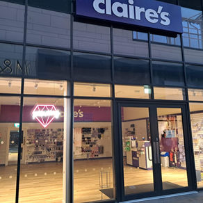 Storefront of Claire's with large glass windows displaying jewelry and accessories. A pink neon diamond light is on inside.