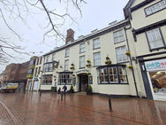 Street view of Swan Hotel, cream facade with flower baskets. Two people walk on wet brick pavement. Overcast sky with bare branches.