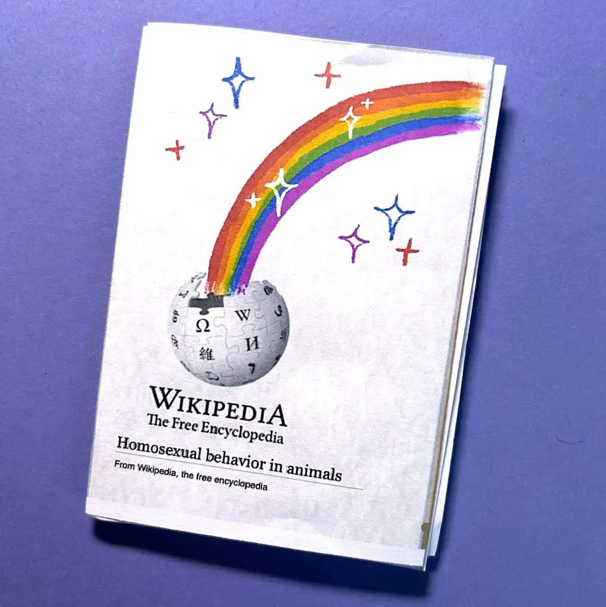 Photograph of a "Homosexual behaviour in animals" mini-zine's front cover, which depicts the Wikipedia logo with a rainbow bursting out of it and the title of the zine beneath. 