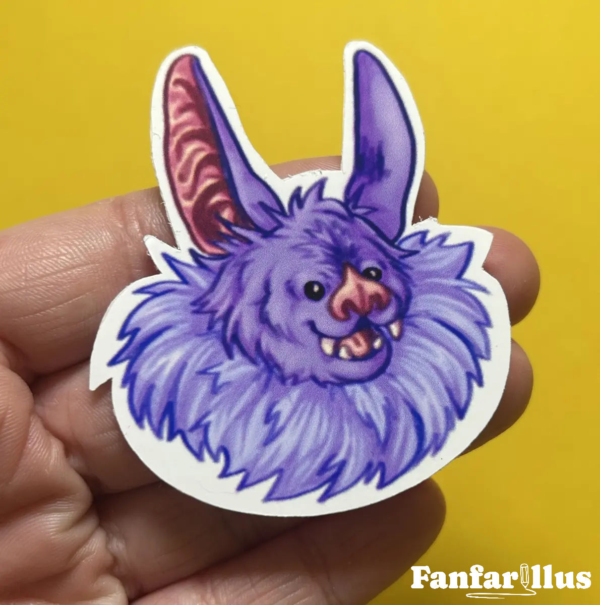 Photograph of a lil bat sticker. The sticker has a glossy finish and depicts a purple, beady eyed bat with an open mouthed smile. The sticker is balanced atop someone's fingers to demonstrate the small size.
