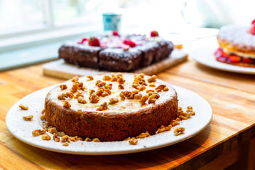  A tempting slice of walnut cake, served at the Endeavour Hotel in Staithes. The cake is presented on a white plate with a dusting of powdered sugar and a sprig of fresh mint on top. The background features a blurred image of a cozy hotel room. The product photography work is credited to Jamie Bulman.