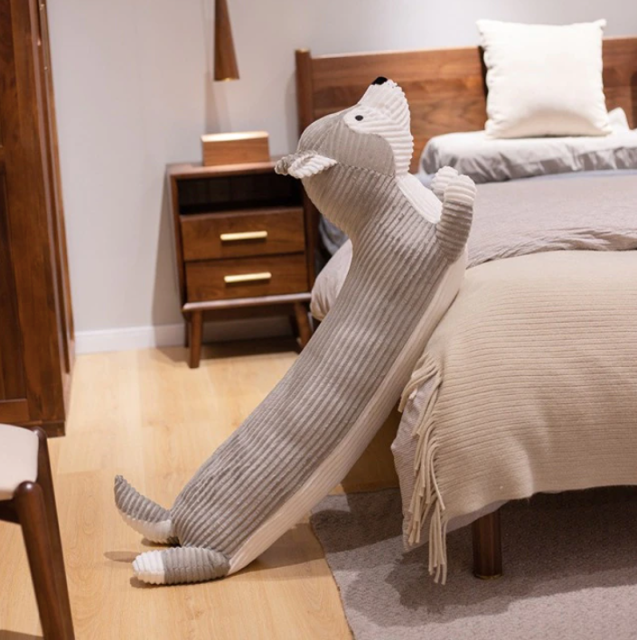 Long gray dog plush pillow leans against a bed; Pawfect Calm–Long Dog Plush Pillow for Rest & Relaxation