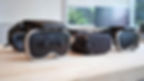 Three VR headsets on a wooden table in a bright room with blurred greenery outside. Black and white design with no visible text.