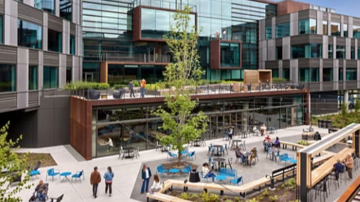 Modern office courtyard with people sitting at blue tables. Glass building facade, greenery, and a tree create a relaxing atmosphere.