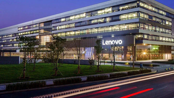 Modern Lenovo building at dusk, large windows illuminated. "Lenovo" sign visible. Street in foreground with light streaks from passing cars.