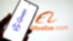 Hand holds smartphone showing "Qwen" logo, with "Alibaba.com" logo in the background. White and orange theme, tech-related setting.