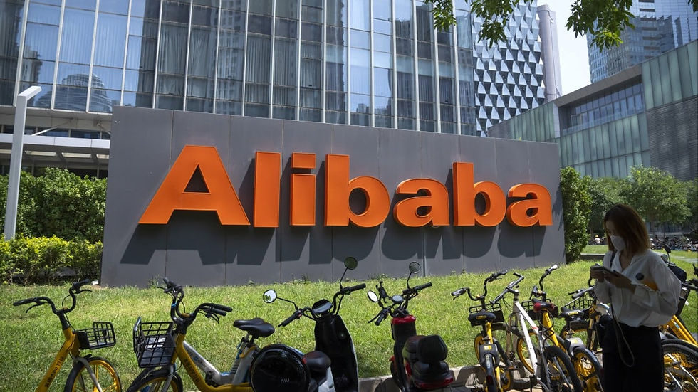 Large "Alibaba" sign in front of a modern glass building. Bicycles in the foreground, a person uses a phone. Bright, greenery visible.