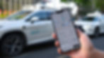 Hand holds a phone showing a map app, with a white autonomous car in the background on a city street. Some Chinese text is visible.