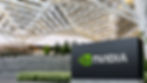 NVIDIA sign in foreground, modern architectural structure and greenery in background. Clear sky visible through geometric roof design.