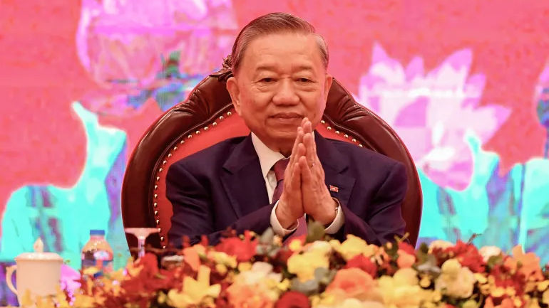Elderly man in a suit sits with hands clasped in front of a floral arrangement on a red, lotus-patterned background, conveying a formal mood.
