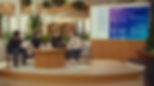 Four people sit around a table with a laptop in a modern office. A large screen displays a tech interface. Green plants and neutral tones.
