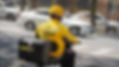 Delivery person on a scooter wears yellow uniform, helmet, and rides on a city street. White car and buildings in background.