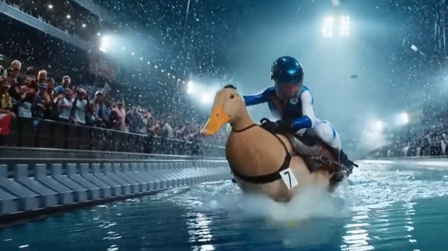 A person in a blue helmet rides a large duck in a water race, surrounded by cheering crowd. Bright floodlights illuminate the scene.