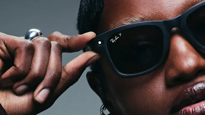Close-up of a person adjusting black Ray-Ban glasses with a camera. Silver ring visible on finger, gray background. Mood is focused.
