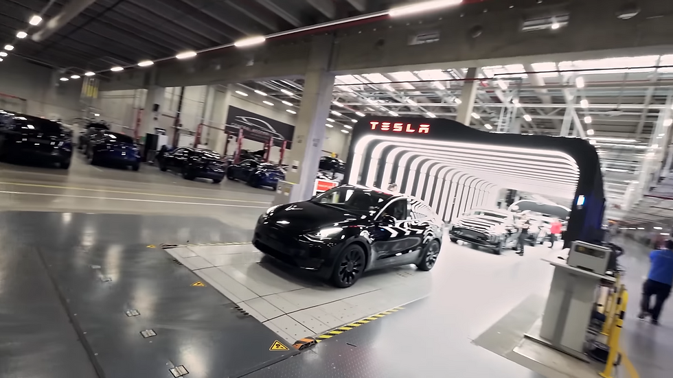 Black Tesla car in a well-lit factory setting, passing through a light tunnel with "Tesla" sign. Other cars and workers in the background.