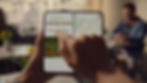 Hands hold a folding phone showing "Adventures in Bloom" webpage and map. Bright indoor café setting with blurred people and flowers.