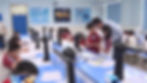 Students in a robotics lab focus on projects, guided by a teacher. Blue tables, robot toys, and posters create a tech-focused setting.