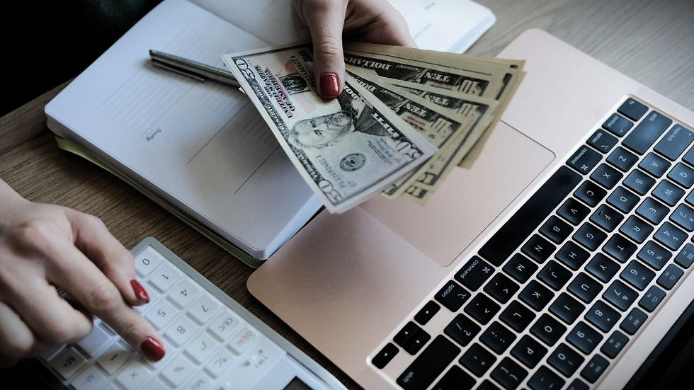Hands holding cash and using calculator next to a laptop and notebook on a wooden desk. The scene suggests a financial task.