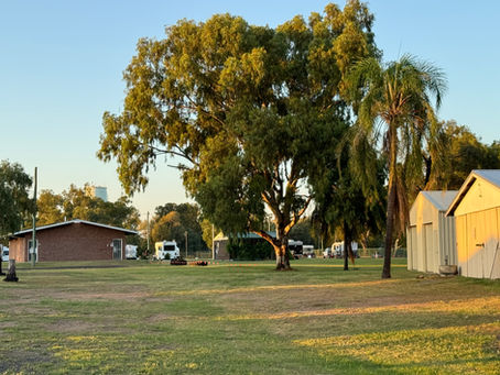 Open grassy field with trees, brick and metal buildings, and parked vehicles under a clear sky at sunset.