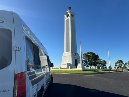 A white camper van parked near a tall white monument on a sunny day. Blue sky, green grass, and trees in the background.