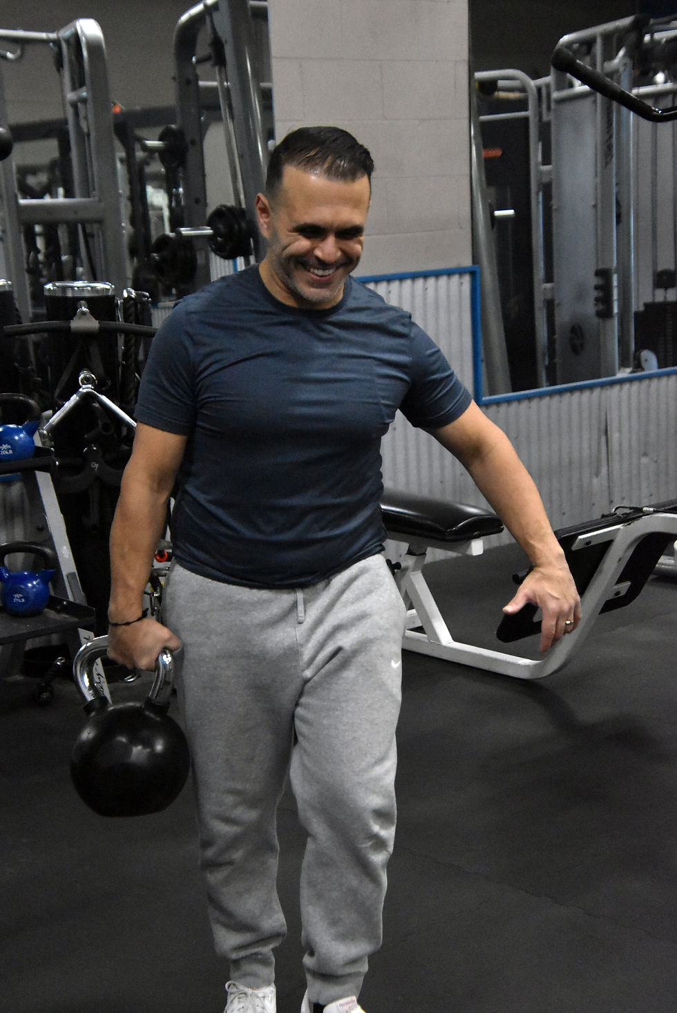 Smiling man in a gym holds a kettlebell beside weight machines, wearing a navy shirt and gray sweatpants.