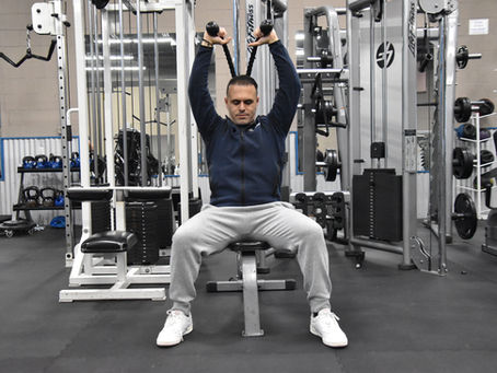 Man in blue jacket and gray pants exercises using a cable machine in a gym. The background includes weight racks and fitness equipment.