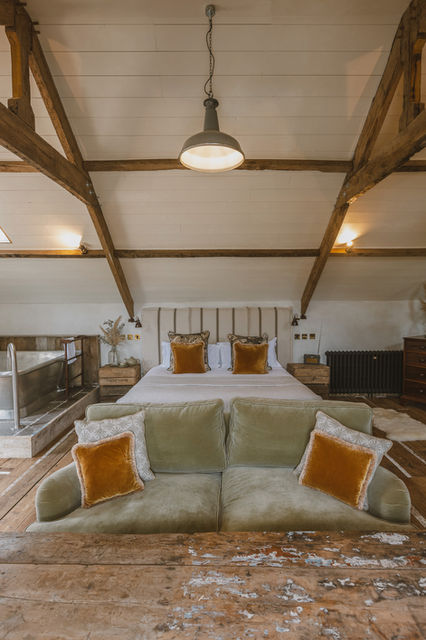 Modern rustic Granary Room at Outbuildings Dorset featuring a velvet green couch, handmade table, bed with wooden framework in the ceiling, and a large Jacuzzi tub in orange and sage green tones