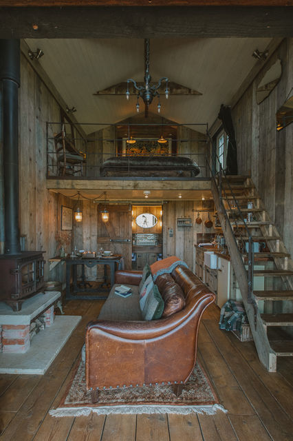Two-story rustic modern Coop Room at Outbuildings Dorset featuring a staircase, bedroom overlooking living and dining area, log burner, leather couch, and wooden interior throughout.