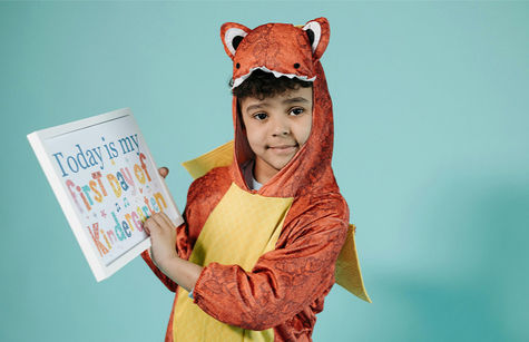 An adorable, elementary school aged boy dressed in a dinosaur costume, smiling and holding a children's book.