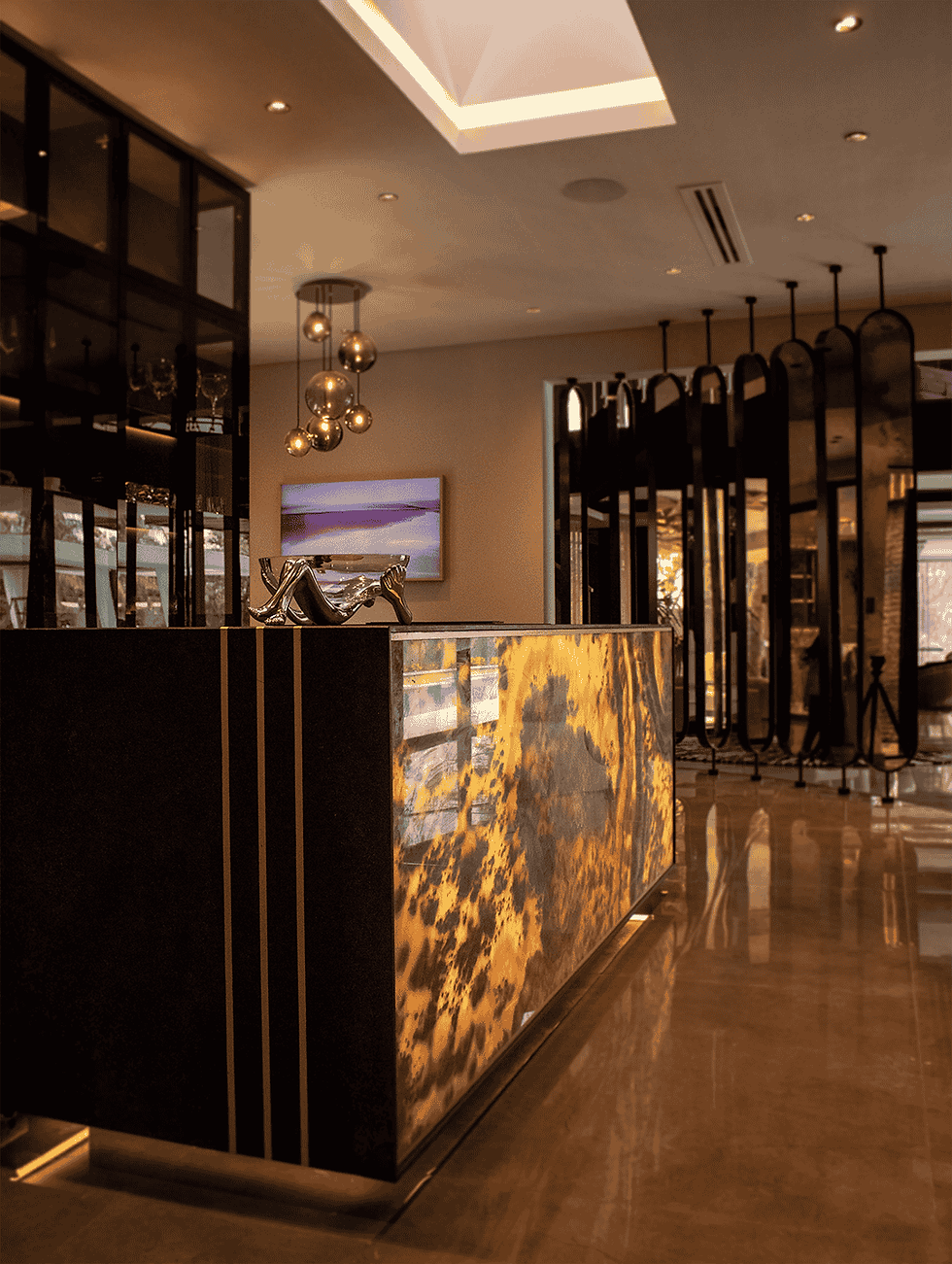 High-end kitchen design showcasing an illuminated island clad in striking onyx stone and brass accents. The space features modern, dark matte-finish cabinets and a textured, backlit splashback in warm taupe tones, creating a sophisticated blend of light and texture.