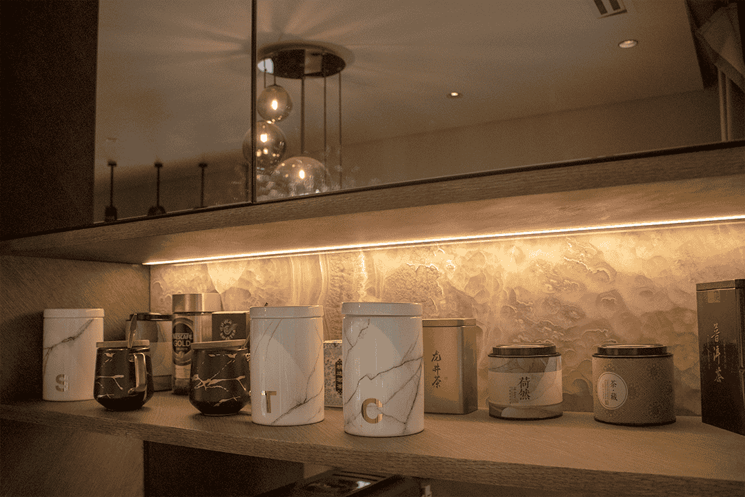 A kitchen splashback with a textured, organic pattern in warm taupe tones, softly backlit to create a glowing effect, contrasts with the sleek matte-finish cabinets and adds a luxurious depth to the modern design.