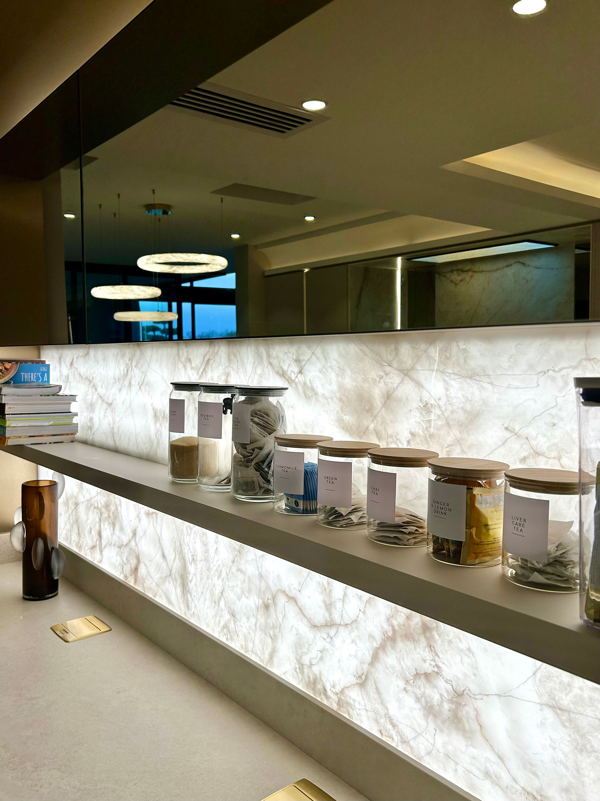 A detailed view of a built-in coffee and tea station featuring a brilliant, backlit white-and-tan onyx stone backsplash that casts a warm, dramatic glow. A minimalist white floating shelf runs across the stone, holding neat glass storage jars with wooden lids and custom labels like "Rooibos Tea" and "Green Tea." Above, a dark mirrored panel reflects the ambient ring pendant lights from across the room.