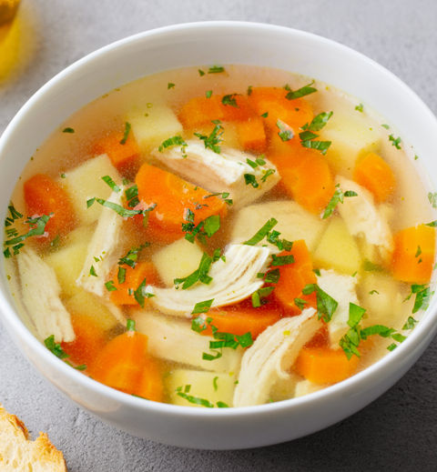 Bowl of chicken soup with carrots and herbs on a gray table. Nearby are a spoon, bread, and a bowl of chopped parsley. Warm, cozy setting.