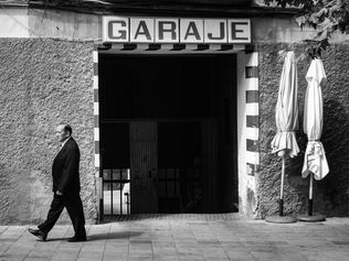 Black and white image of a man in a suit walking past a garage entrance. Two closed umbrellas stand to the right, evoking a serene, urban scene.