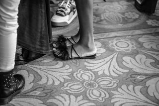 Black and white image of people standing on a patterned carpet. Visible footwear includes decorative sandals, sneakers, and studded boots, conveying a casual and stylish atmosphere.