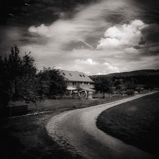 Black and white photo of a farmhouse on a rural path, surrounded by trees and fields under a dramatic, cloud-filled sky. Mood is serene and nostalgic.
