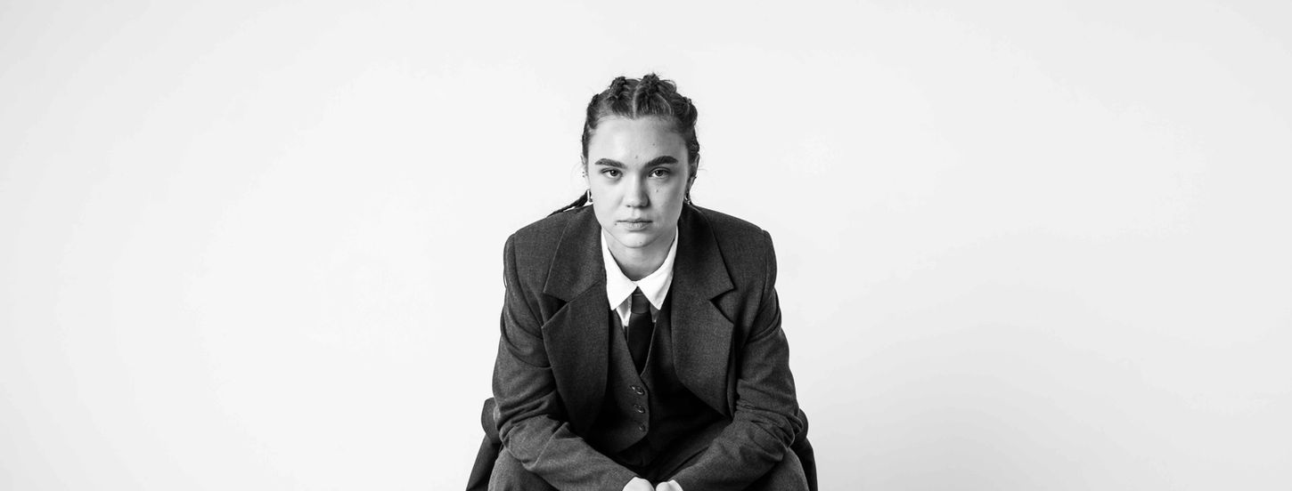 Woman in suit seated on stool, facing forward.