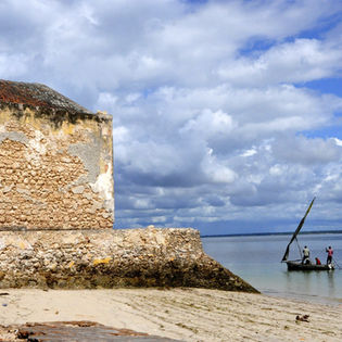Old building and boat on water with people under cloudy sky.