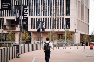 Person with a backpack walking towards a modern building adorned with "UCL" banners. Sparse trees and poles line the pathway, suggesting a campus setting.