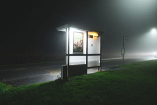 A bus stop shelter illuminated in foggy conditions at night, with a blank advertisement space on the side.