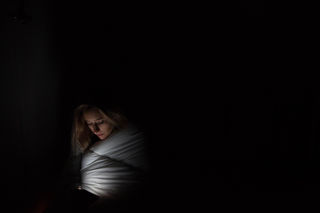 A person wrapped in a blanket looks at a phone in a dimly lit room, creating a tranquil and intimate atmosphere. The phone light softly illuminates their face.
