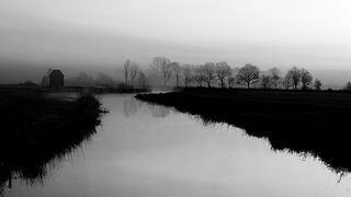 A misty landscape in black and white shows a quiet river with bare trees and a solitary house on the left. The scene is serene and contemplative.
