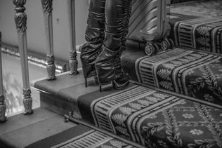 Black and white image of a person in high-heeled boots climbing ornate carpeted stairs, accompanied by a suitcase, suggesting travel.