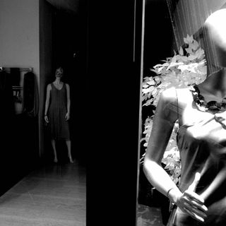 Black and white image of two mannequins. The foreground mannequin wears a dress and necklace, illuminated next to leaves. The background mannequin stands in shadow, creating an eerie contrast.